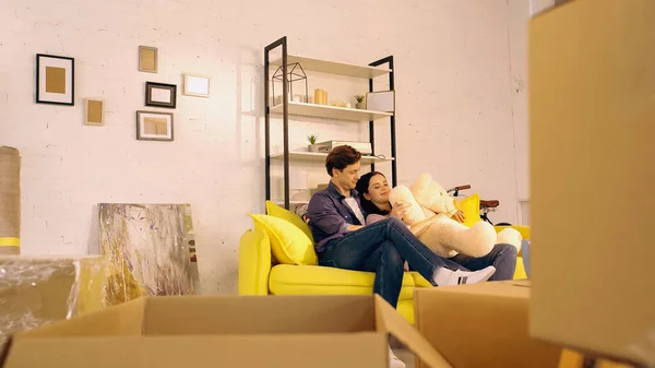 Felice coppia di relax sul divano con orsacchiotto vicino scatole in nuova casa — Foto stock