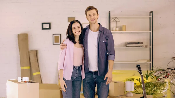 Cheerful couple standing and hugging near boxes in new home — Stock Photo