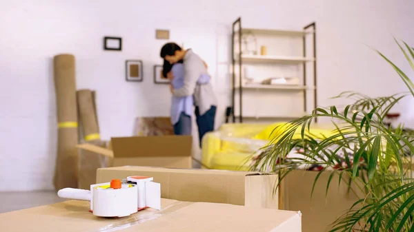 Fita adesiva em caixas perto de casal borrado abraçando em nova casa — Fotografia de Stock