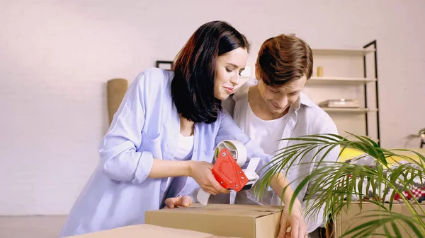Cheerful couple using scotch tape while packing box — Stock Photo