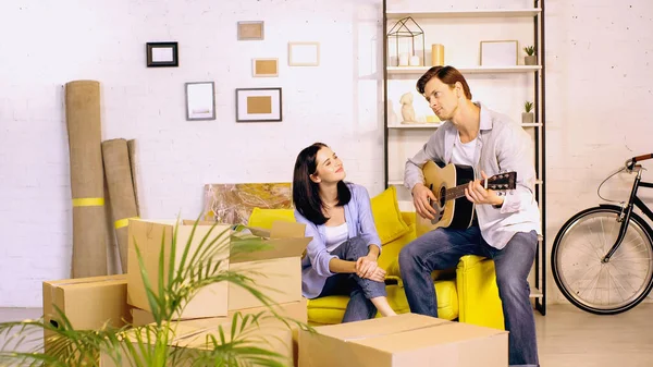 Man playing acoustic guitar near happy girlfriend sitting on couch in new home — Stock Photo