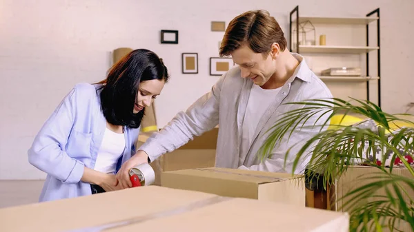 Joyful couple using scotch tape while packing box — Stock Photo