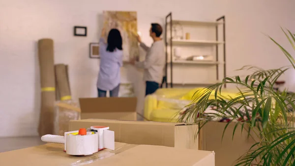 Scotch tape on boxes near blurred couple hanging painting in new home — Stock Photo