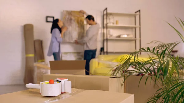 Scotch tape on carton boxes near blurred couple hanging painting in new home — Stock Photo