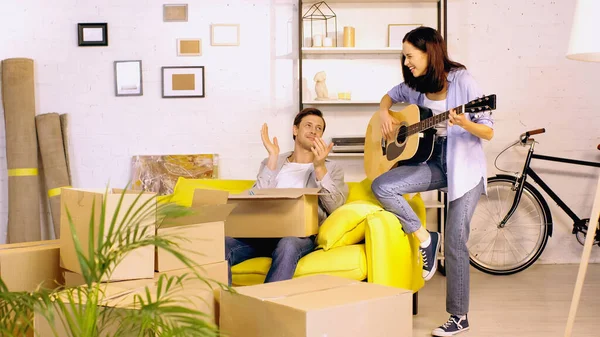 Jeune femme jouer de la guitare près de copain applaudissant dans nouvelle maison — Photo de stock