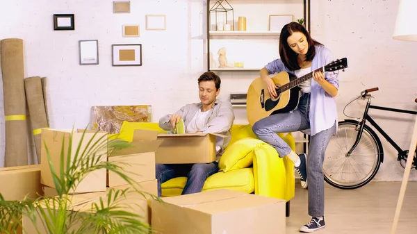 Jeune femme jouant de la guitare près du petit ami déballer la boîte dans une nouvelle maison — Photo de stock