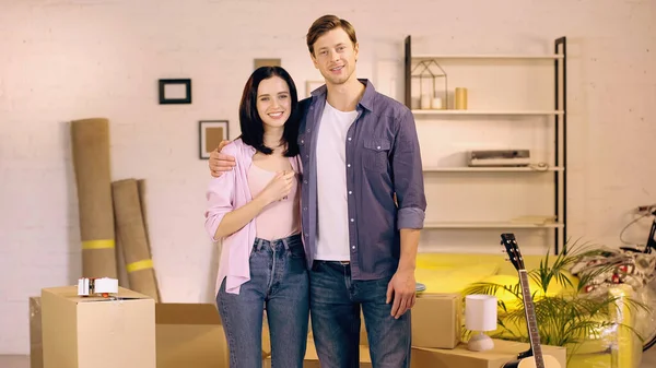 Feliz pareja sonriendo y de pie cerca de cajas en un nuevo hogar - foto de stock