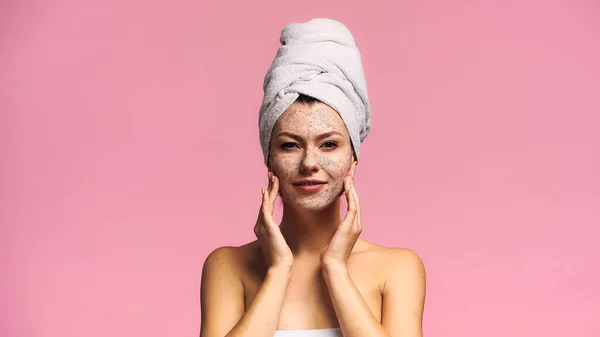 Mujer sonriente con toalla de rizo en la cabeza aplicando exfoliante en la cara aislado en rosa - foto de stock