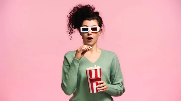 Amazed woman in 3d glasses holding bucket of popcorn isolated on pink — Stock Photo