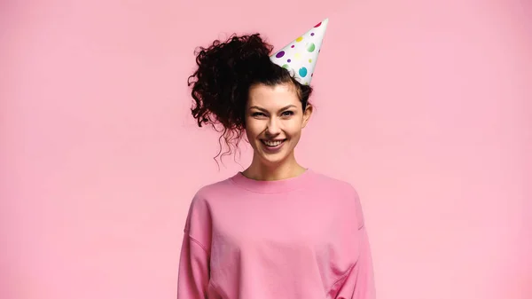 Fröhlich lockige Frau mit Partymütze lächelt in die Kamera isoliert auf rosa — Stockfoto