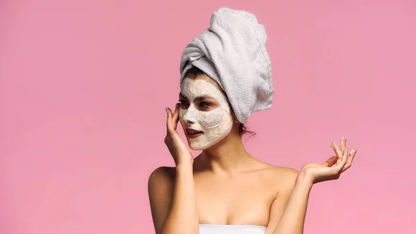 Mujer joven aplicando mascarilla de arcilla en la cara mientras mira hacia otro lado aislado en rosa - foto de stock