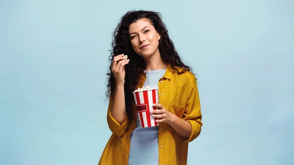 Happy woman with bucket of popcorn looking at camera isolated on blue — Stock Photo