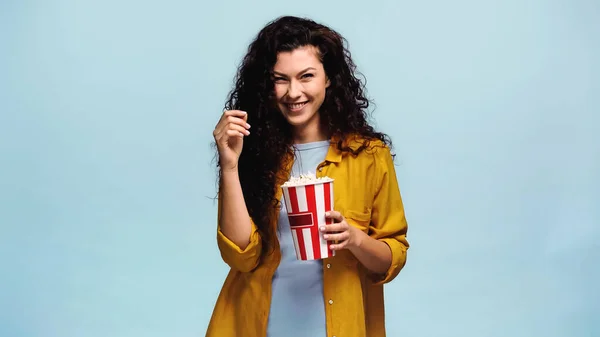 Mujer excitada con cubo rayado de palomitas de maíz sonriendo a la cámara aislada en azul - foto de stock