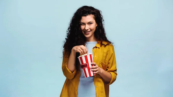 Cheerful woman eating popcorn and smiling a camera isolated on blue — Stock Photo