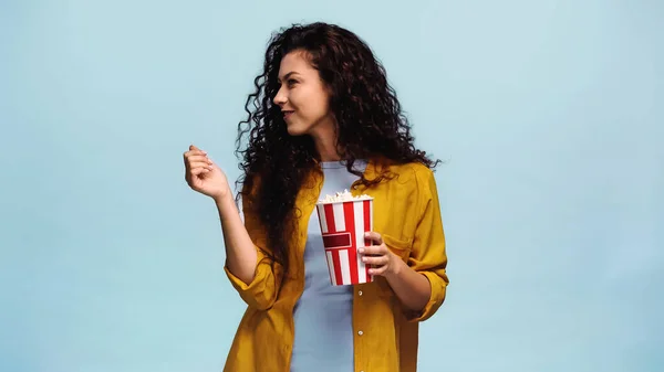 Curly woman with popcorn smiling while looking away isolated on blue — Stock Photo