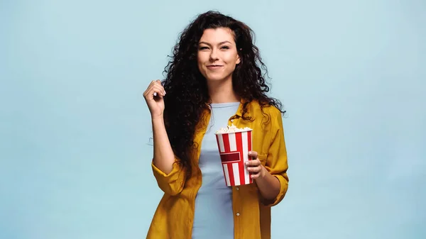 Zufriedene Frau blickt in die Kamera, während sie Popcorn isoliert auf blauem Grund hält — Stockfoto