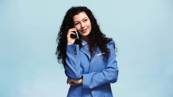 Morena mujer mirando hacia otro lado y sonriendo mientras habla en el teléfono celular aislado en azul - foto de stock