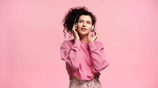 Feliz joven mujer ajustando auriculares mientras escucha música aislada en rosa - foto de stock