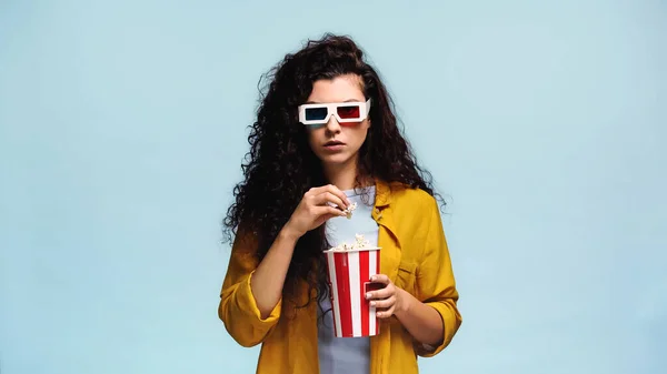 Curly brunette woman in 3d glasses holding striped bucket with popcorn isolated on blue — Stock Photo