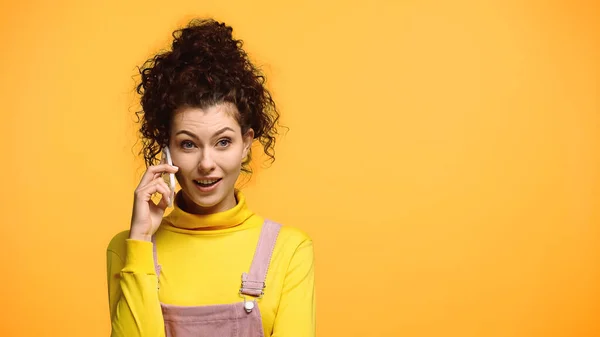 Mujer sorprendida en cuello alto amarillo hablando en teléfono móvil aislado en naranja - foto de stock