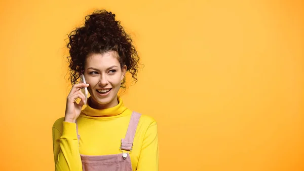 Mujer alegre con el pelo ondulado hablando en el teléfono inteligente aislado en naranja - foto de stock
