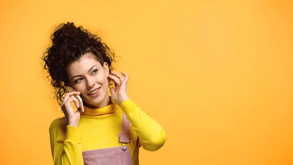 Mujer feliz hablando en el teléfono móvil mientras toca el pelo aislado en naranja - foto de stock
