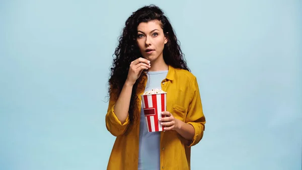 Surprised woman in orange shirt eating popcorn isolated on blue — Stock Photo