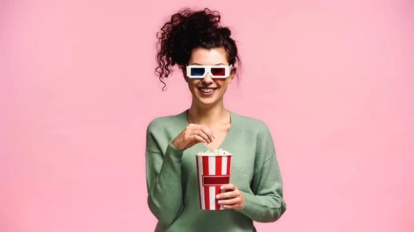 Fröhliche Frau in grünem Pullover und 3D-Brille, die Popcorn isoliert auf rosa isst — Stockfoto