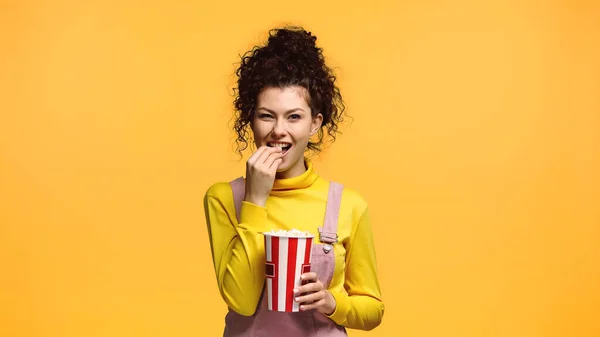 Mujer alegre con el pelo ondulado comer palomitas de maíz aislado en naranja - foto de stock