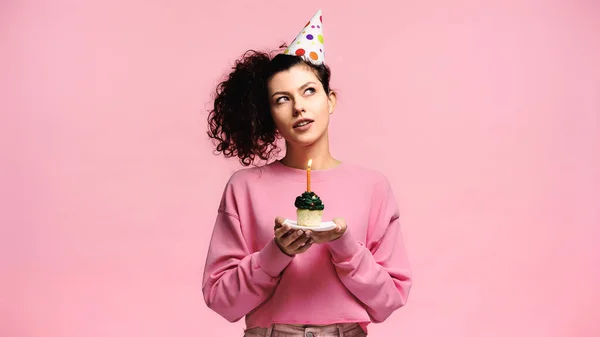 Мрійлива жінка робить бажання, тримаючи кекс на день народження ізольовано на рожевому — стокове фото