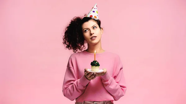 Вдумчивая женщина с праздничным кексом загадывает желание, изолированное на розовый — стоковое фото