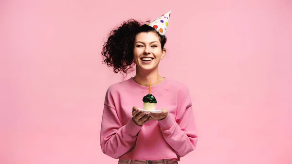 Весела жінка у вечірній кепці, що тримає кекс на день народження зі свічкою ізольовано на рожевому — стокове фото