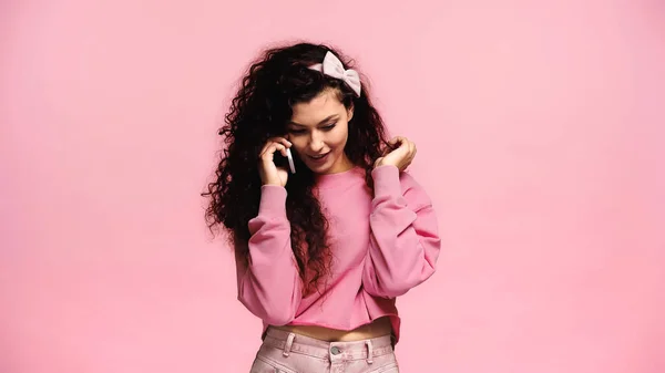 Mujer bonita y positiva tocando el cabello mientras habla en el teléfono celular aislado en rosa - foto de stock