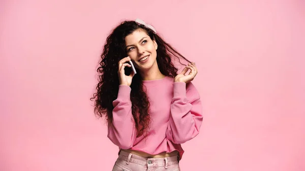 Mujer alegre tocando el cabello y mirando hacia otro lado mientras habla en el teléfono inteligente aislado en rosa - foto de stock