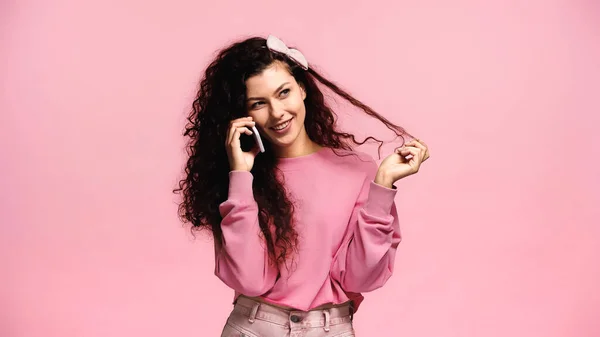 Mujer complacida tocando el pelo mientras habla en el teléfono celular aislado en rosa - foto de stock