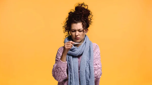 Unzufriedene Frau in Strickpullover und Schal blickt auf Thermometer in Orange — Stockfoto