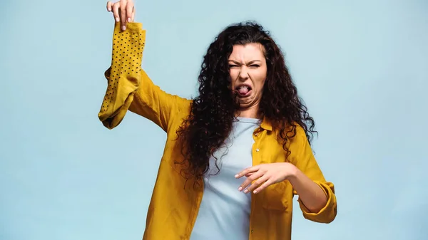 Curly woman grimacing and sticking out tongue while holding smelly socks isolated on blue — Stock Photo