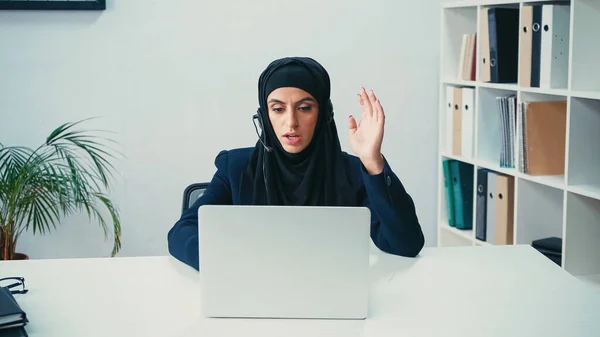 Muslimische Frau gestikuliert mit Kopfhörer, während sie Laptop in Callcenter benutzt — Stockfoto