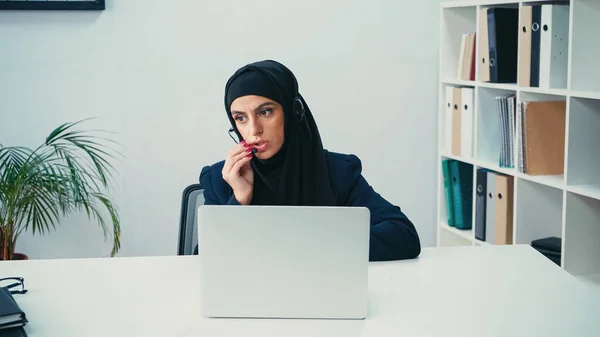 Mujer de negocios musulmana en auriculares usando micrófono cerca de la computadora portátil en el centro de llamadas - foto de stock