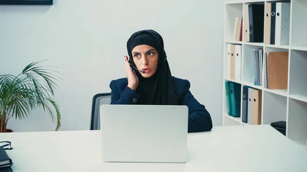 Femme musulmane dans un casque utilisant un microphone et parlant près d'un ordinateur portable dans un centre d'appel — Photo de stock