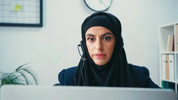 Femme musulmane dans le hijab en utilisant un casque et en regardant un ordinateur portable flou dans le centre d'appel — Photo de stock