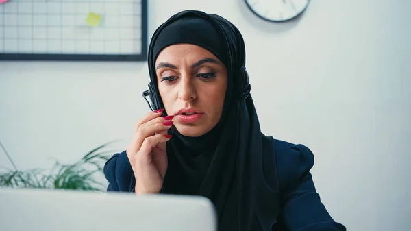 Muslimische junge Frau im Hidschab benutzt Kopfhörer in Nähe eines verschwommenen Laptops im Callcenter — Stockfoto