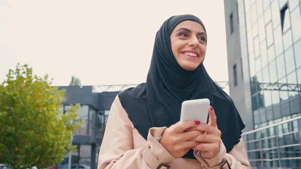 Happy young muslim woman in hijab messaging on smartphone outside — Stock Photo