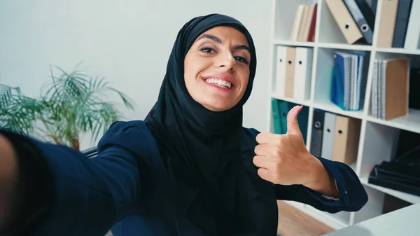 Счастливая мусульманка-бизнесвумен, показывающая большой палец, делая селфи — стоковое фото
