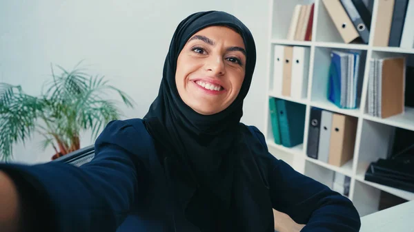 Cheerful muslim businesswoman looking at camera while taking selfie — Stock Photo