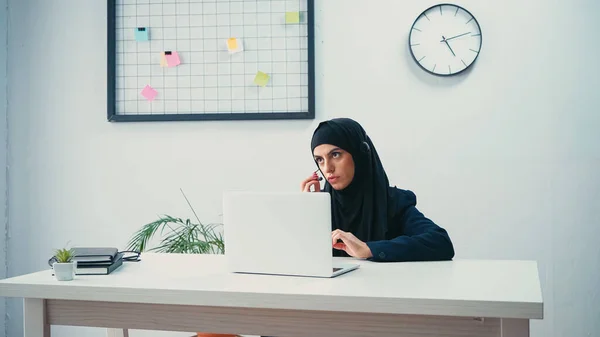 Donna musulmana in hijab e auricolare regolazione microfono vicino laptop in call center — Foto stock