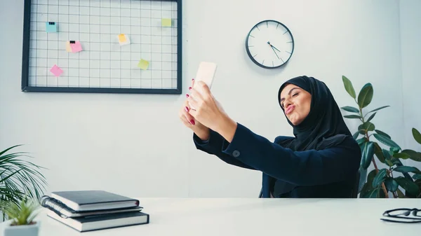 Muslim businesswoman pouting lips and taking selfie on smartphone in office — Stock Photo