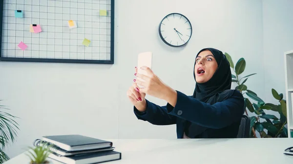 Мусульманская предпринимательница с открытым ртом делает селфи на смартфоне в офисе — стоковое фото