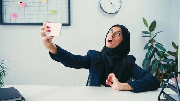 Femme d'affaires musulmane à bouche ouverte prenant selfie sur téléphone portable dans le bureau — Photo de stock