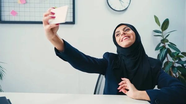 Femme d'affaires musulmane et heureuse prenant selfie sur smartphone au bureau — Photo de stock